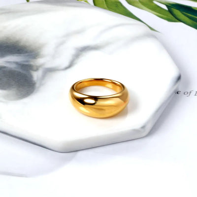 A polished 18K gold-plated stainless steel low dome band with a 9.7mm width sitting on marble coaster. A perfect minimalist ring for stacking or solo wear.