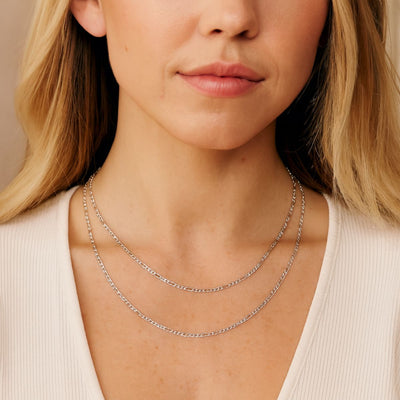 Close-up of a woman with blonde hair wearing three Sterling Silver 2.5mm 3+1 Figaro chains layered at 40cm and 50cm lengths. The polished chains sit neatly at the collarbone and neckline, styled with a light V-neck top.