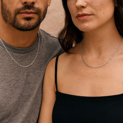 A man and woman wearing matching 55cm long, 2mm wide 3+1 Figaro chains in Sterling Silver. The polished chains rest neatly at the collarbone, styled with casual tops against a soft neutral background.