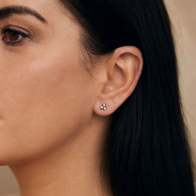Close-up of a woman wearing 3mm sterling silver three-ball stud earrings. The polished spheres form a neat triangular shape, positioned on her earlobe with dark hair tucked behind her ear.