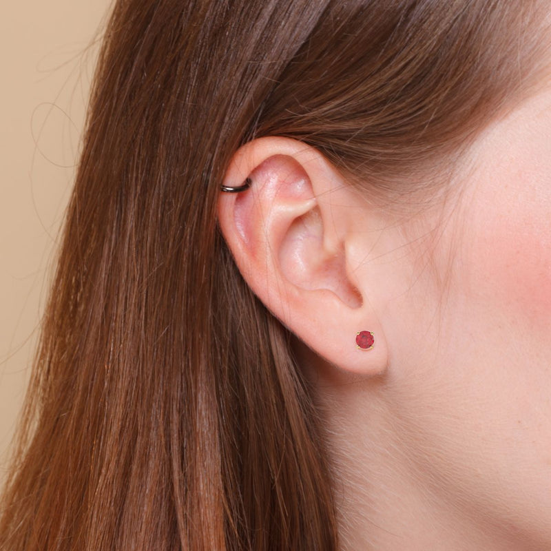 In this elegant photo, a model gracefully wears a 4mm natural ruby earring in her right ear, with a sleeper on the top of her ear for a modern and classy style. Shop gold stud earrings from Jewels of St Leon.