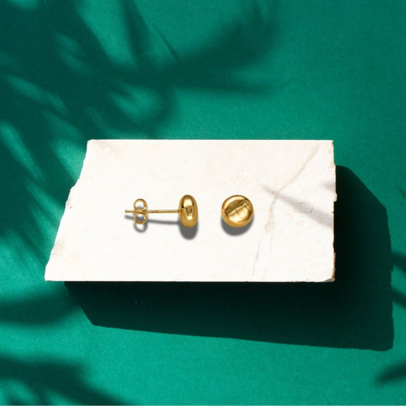 The 6mm Heavy Flat Ball Earrings in 9ct Gold are artfully displayed on a rock with a lush green background, emphasizing their radiant shine and natural charm.