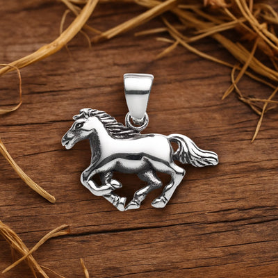 Sterling silver horse pendant resting on rustic timber floorboards with scattered hay. The pendant catches soft natural light, highlighting its polished finish and detailed craftsmanship in a warm, rural-inspired setting.