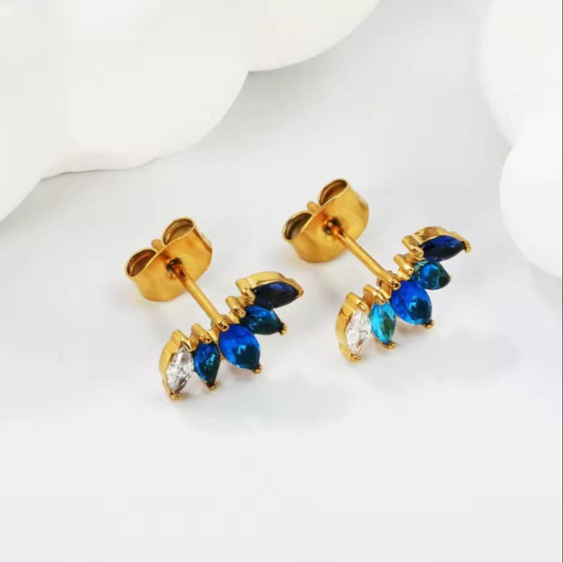 Angled view of tiny ombré blue and clear CZ fan studs on a white surface, displaying gold-plated posts, butterfly backs and vivid marquise stones.