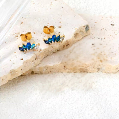Lifestyle shot of petite 12 × 7 mm fan-shaped stud earrings with ombré sapphire-blue and clear marquise CZs in 18 K gold-plated stainless steel, resting on natural limestone.