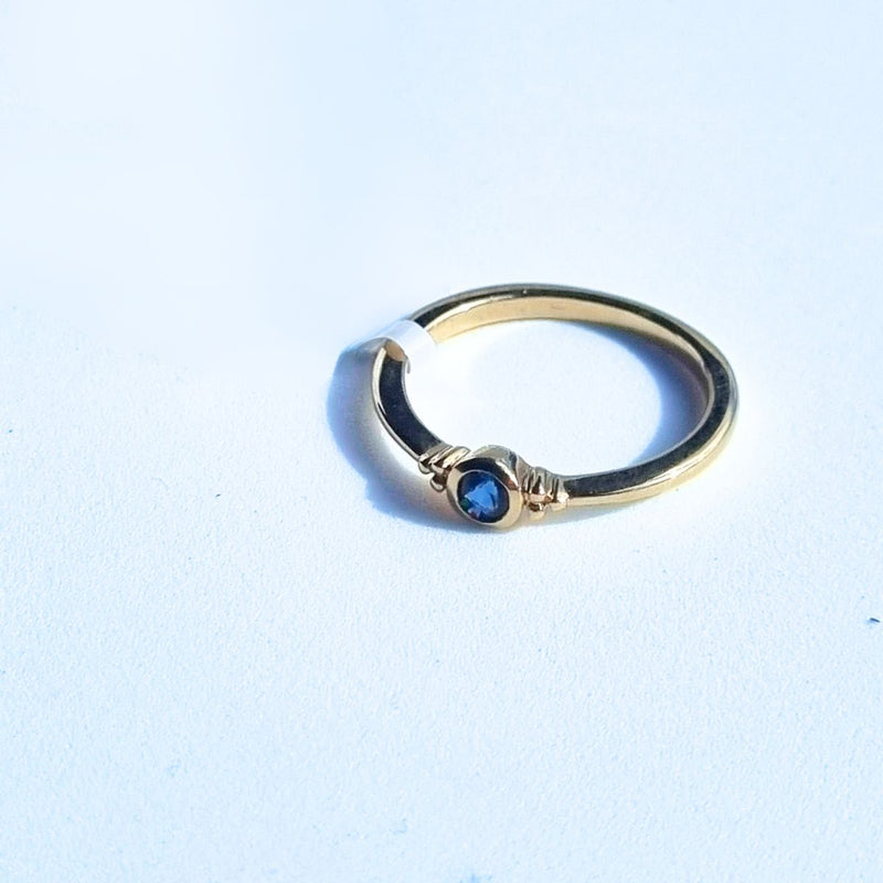 The ELLIN Ring lying flat at a 45° angle to the left, emphasising the sleek and minimalist band of 9kt yellow gold and the exquisite blue sapphire, demonstrating its elegant and stackable design.