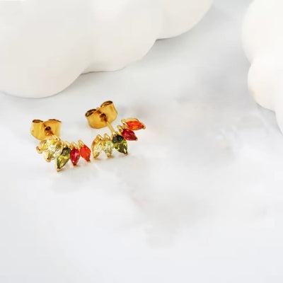 Close-up of tiny fan-shaped stud earrings with marquise CZs in yellow, olive, pink and orange on a white backdrop beside cloud-shaped décor.