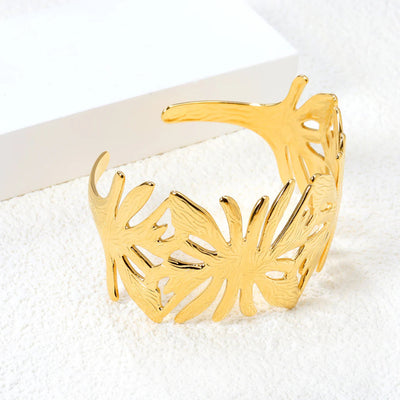 A statement leaf-inspired cuff bangle with an open wrap-around design sits on a soft carpet. It is beautifully polished in 18K gold-plated stainless steel for a luxurious finish. Get the look of high fashion for an affordable price.