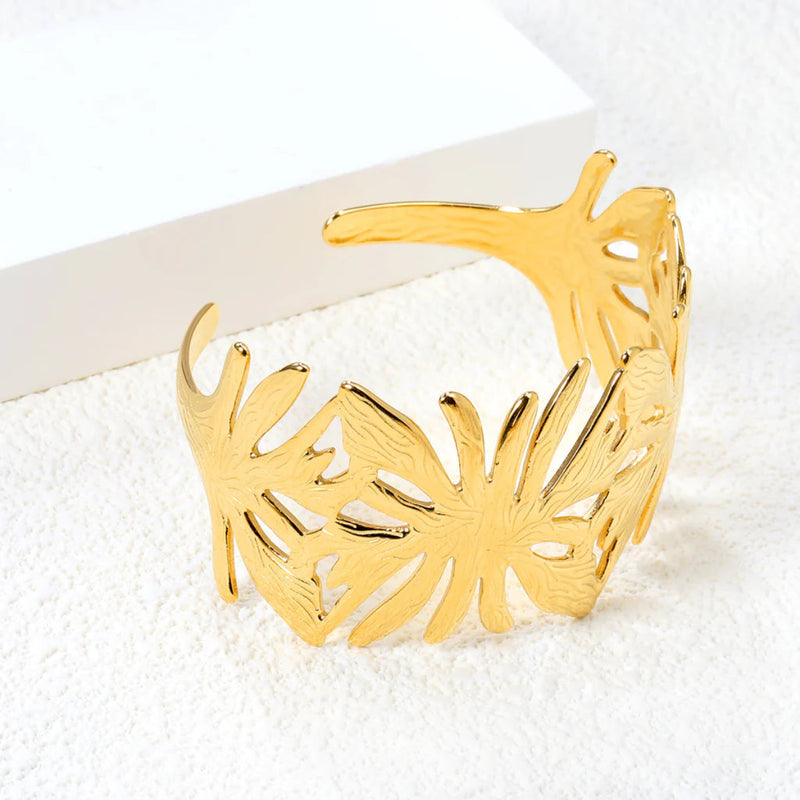 A statement leaf-inspired cuff bangle with an open wrap-around design sits on a soft carpet. It is beautifully polished in 18K gold-plated stainless steel for a luxurious finish. Get the look of high fashion for an affordable price.
