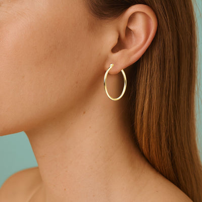 A female model with chestnut coloured hair, wearing 25mm x 2mm 14ct Gold Plated Sterling Silver Hoop Earrings.