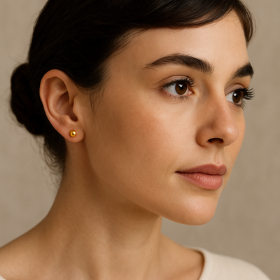 Close-up portrait of a brunette woman looking to her left, wearing a 5 mm gold flat ball stud earring in her right ear against a soft neutral background. Shop now at Jewels of St Leon.