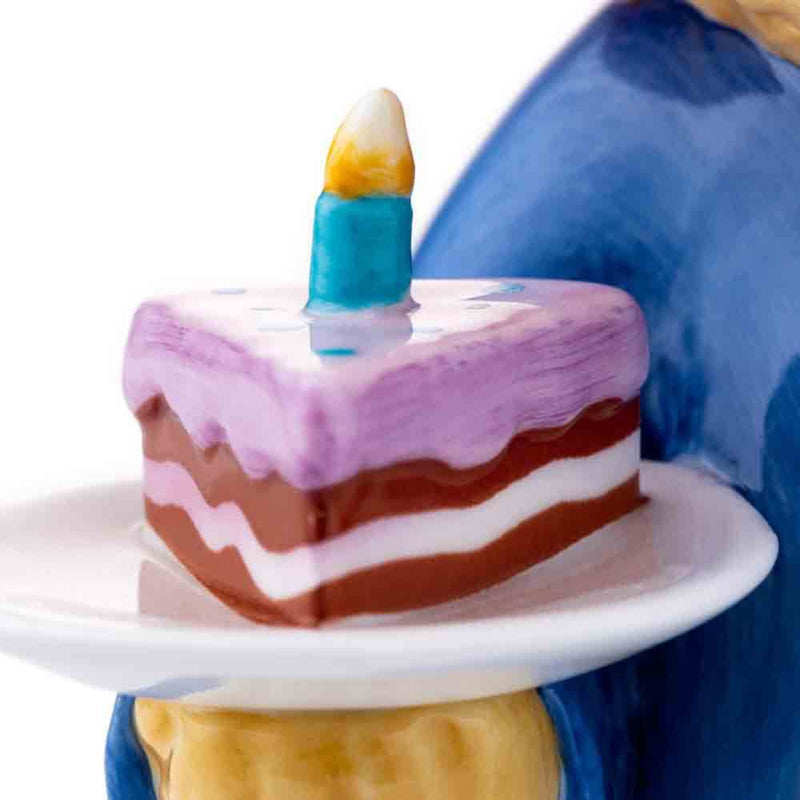 Close up shot of a slice of a three layer chocolate cake with strawberry frosting and a single candle being held by Paddington.