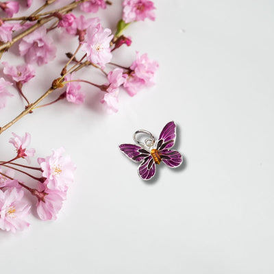 The Purple Enamel Butterfly Charm Pendant displayed against a soft grey background, framed by delicate flowers that complement its vibrant hues and elegant design.