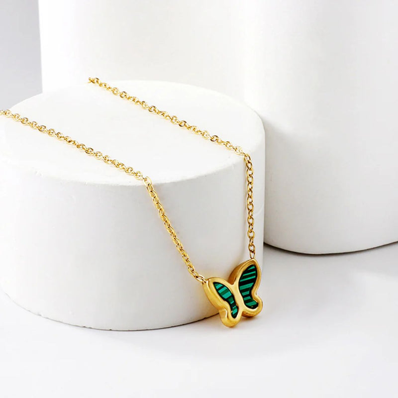 The Green Butterfly Necklace hanging from the side of a round display, capturing the vibrant green enamel wings and delicate gold chain against a soft backdrop.