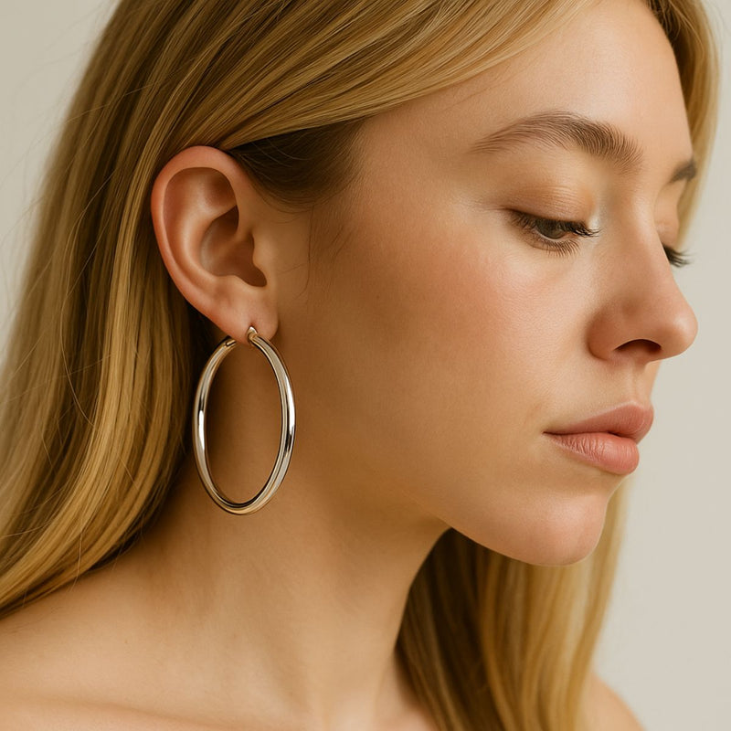 Close-up of a woman wearing 2mm x 50mm Sterling Silver hoop earrings. The polished hoops sit just below her jawline, highlighting their large size and lightweight tubular design. Her blonde hair is tucked back, and the background is softly blurred.