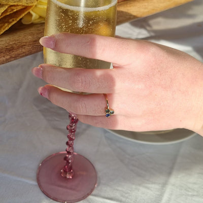 Lifestyle Shot: The Katrine Ring shines brightly on a model's hand as she holds a glass of champagne on the Gold Coast. The green, teal, and blue sapphires glisten in the sunlight, adding elegance to any celebration.