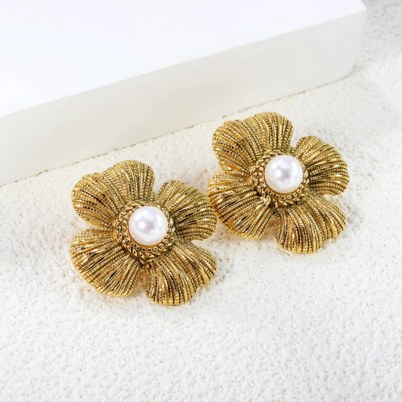 Pair of textured gold-plated four-petal flower stud earrings with pearl centres, styled on a white surface beside sculptural ceramic shapes.